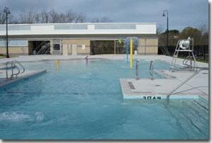 A swimming pool with fountains in the shallow area and a building on the far side for lockers